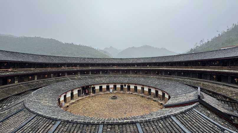 Interior Tulou Hakka di Meizhou — rumah lingkar bertingkat tiga dengan koridor kayu dan halaman tengah, dikelilingi pegunungan berkabut